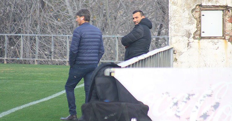 Alfonso Serrano, de espaldas, informando a Raúl Agné de los últimos movimientos del mercado con la imagen de la denostada Ciudad Deportiva visible en el abandono del muro y la valla de publicidad estática.