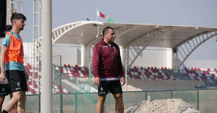 Germán Crespo, siguiendo las evoluciones del entrenamiento, con Javi Flores a la izquierda y al fondo una grada con público siguiendo un partido en las instalaciones de la Federación de Fútbol de Baréin.