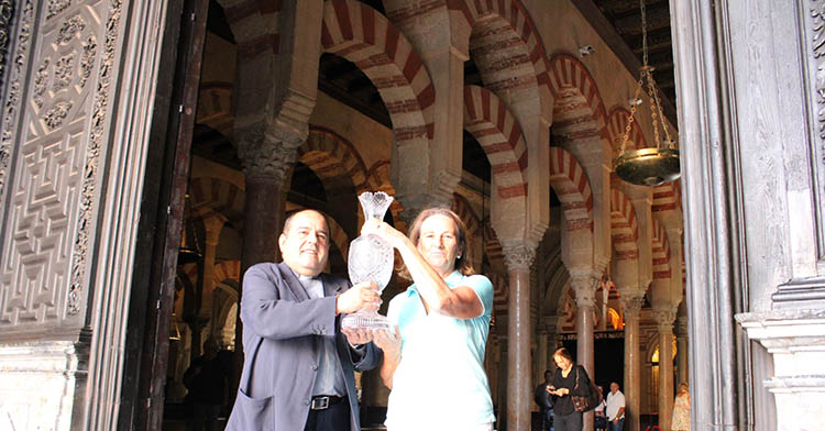 El canónigo del Cabildo Catedral de Córdoba, José Juan Jiménez Güeto, sostiene junto a María del Mar Romero, presidenta del Real Club de Campo, el trofeo de la Solheim Cup ante el bosque de columnas de la Mezquita Catedral al fondo.